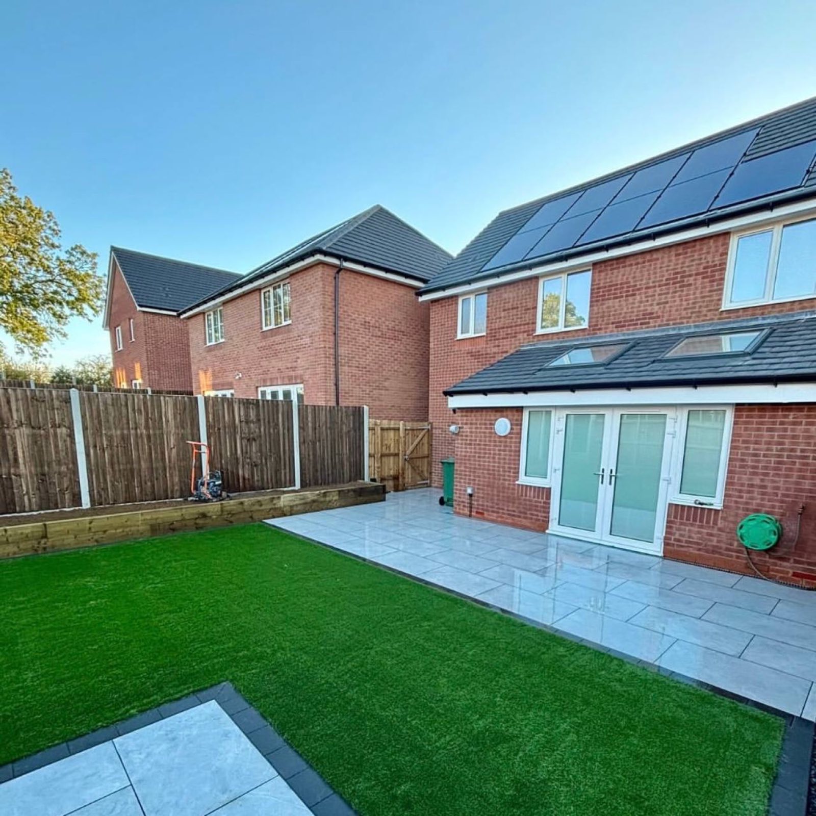 A bare new-build plot turned into a usable garden: light porcelain terrace off the French doors, artificial grass to a dark sett edge, and timber sleeper edging against the fence line.